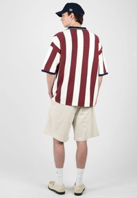 Homme portant une casquette marine, une chemise à manches courtes rayée verticalement bordeaux et blanche, un short beige, des chaussettes blanches et des chaussures beiges, debout de dos.