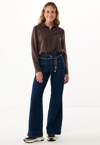 Camicia marrone a maniche lunghe con colletto abbinata a jeans a gamba larga blu scuro, caratterizzati da una cintura con catena e una vestibilità rilassata.