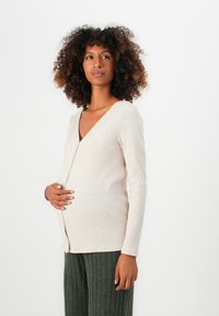 Beige ribbad cardigan med V-ringning och knappar, bärs över gröna ribbade byxor. Cardiganen har långa ärmar och en avslappnad passform.