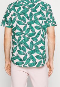 Uomo che indossa una camicia a maniche corte con motivo di foglie tropicali verdi e pantaloni rosa chiaro, mostrato di spalle su sfondo bianco.