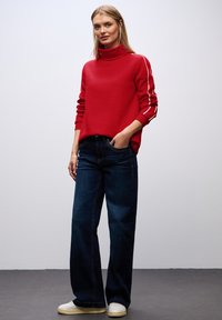 Suéter rojo de cuello alto con rayas blancas en los laterales, combinado con jeans de mezclilla oscura de pierna ancha. Modelo con zapatillas claras, de pie contra un fondo liso.
