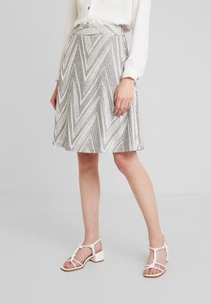 Jupe à motif chevron gris et blanc en tissu texturé, longueur genou, avec une taille haute. Associée à des sandales à talons blanches à brides.