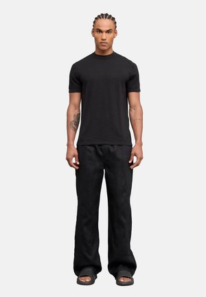 Uomo con i capelli intrecciati che indossa una maglietta nera aderente a maniche corte, pantaloni neri larghi e sandali neri, in piedi su uno sfondo chiaro e uniforme.