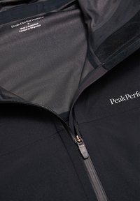 Giacca nera Peak Performance con cerniera frontale parzialmente aperta, che mostra l'etichetta interna del colletto con la scritta "Made in Bangladesh" in testo bianco.