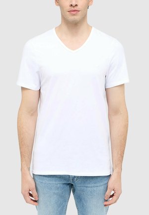 Junger Mann trägt ein schlichtes weißes V-Ausschnitt Kurzarm-T-Shirt und hellblaue Jeans, steht vor neutralem Hintergrund.