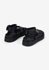 Sandali platform neri con ampie cinghie incrociate e fibbia sul retro. Realizzati in materiale liscio, presentano una suola piatta con una superficie strutturata.