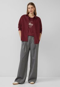 Chemise rouge bourgogne à boutons superposée sur un t-shirt graphique bordeaux, associée à un pantalon large rayé gris et des chaussures à enfiler beiges.