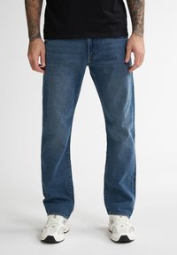Blauwe spijkerbroek met een rechte pasvorm, voorzien van een klassieke vijf-pocketsontwerp en lichte vervaging, gecombineerd met witte sneakers.