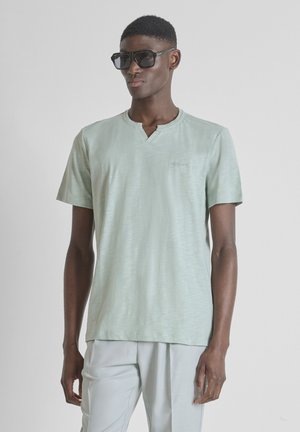 Joven con gafas de sol negras sobredimensionadas, camisa de manga corta verde menta clara y pantalones gris claro sobre fondo liso.