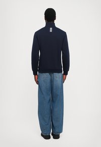 Marineblå crew neck sweatshirt med høy krage og hvite striper på baksiden, kombinert med lyseblå denimjeans med vide ben og svarte sko.