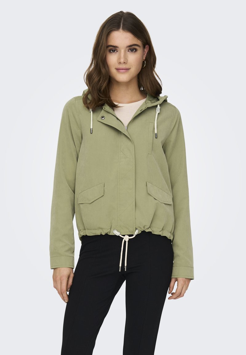 Veste coupe-vent vert olive clair avec un ourlet à cordon, des poches avant et une capuche, dotée d'une fermeture zip et pression. Texture de tissu lisse.