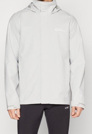Mann trägt eine hellgraue wasserdichte Jacke mit Kapuze, Reißverschluss und Klettverschlüssen an den Bündchen, kombiniert mit schwarzen Sporthosen.