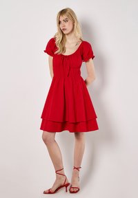 Rode, gelaagde jurk met korte puffmouwen, vierkante halslijn, trekkoorddetails en gestructureerde stof. Gepaarde met rode, strapsandalen.
