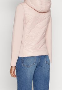 Ljusrosa jacka med ett kviltat puffer-design på baksidan, utrustad med huva och kombinerad med blå denimjeans. Släta och texturerade material.