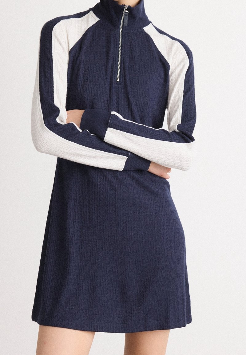 Femme portant une robe en maille bleu marine et blanche à manches longues, avec une fermeture à glissière sur le devant et une texture côtelée, les bras croisés à la taille.