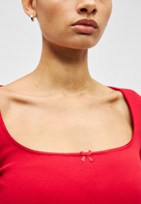 Crveni rebrasti top s kvadratnim izrezom, ukrašen malom odgovarajućom mašnom i dekorativnim rubom. Meka tekstura i kroj prilagođen tijelu.