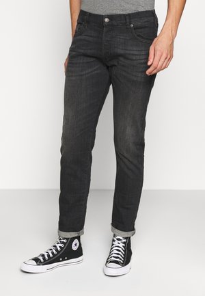 Homme portant un jean gris foncé retroussé et des baskets montantes noires Converse, debout devant un fond uni.