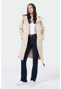 Beige pufferjack met een doorgestikt ontwerp, capuchon en zwarte accenten, gedragen over een witte top en donkere jeans met een flared pasvorm. Witte sneakers.