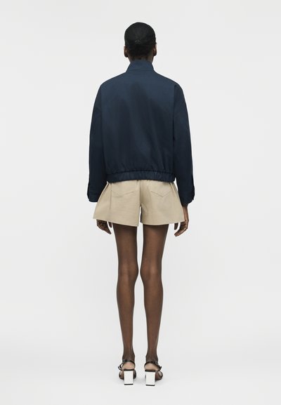 Άτομο που στέκεται με την πλάτη προς τα εμπρός, φοράει σκούρο μπλε μπουφάν με ελαστική μέση, μπεζ σορτς με πίσω τσέπες και μαύρα σανδάλια με λευκές χοντρές γόβες.