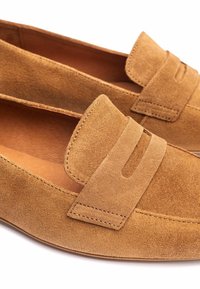 Lichte suède loafers met een platte zool en een gestikte accentband aan de bovenzijde. De textuur is zacht en glad, met minimale details.