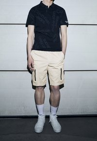 Černé polo tričko s opakovaným textem "Calvin Klein Jeans", spárované se světle béžovými cargo šortkami s černými zipovými kapsami a bílými teniskami.