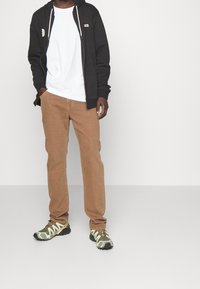 Mann steht mit teilweise in den Taschen, trägt eine schwarze Zip-Jacke, ein weißes T-Shirt, braune Hose und grün-graue Wanderschuhe vor einem hellen Hintergrund.