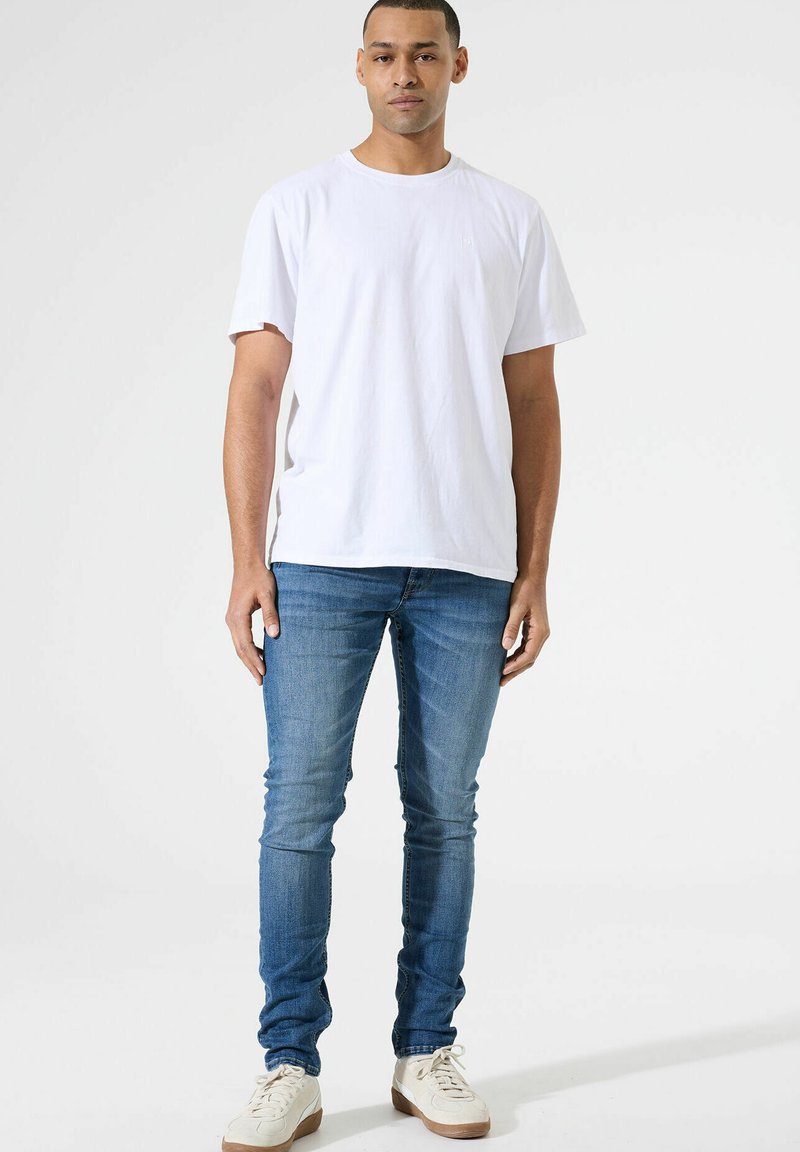 Witte katoenen T-shirt met korte mouwen, gecombineerd met lichtblauwe denim jeans. Model draagt off-white sneakers. Eenvoudig ontwerp, geen patronen.