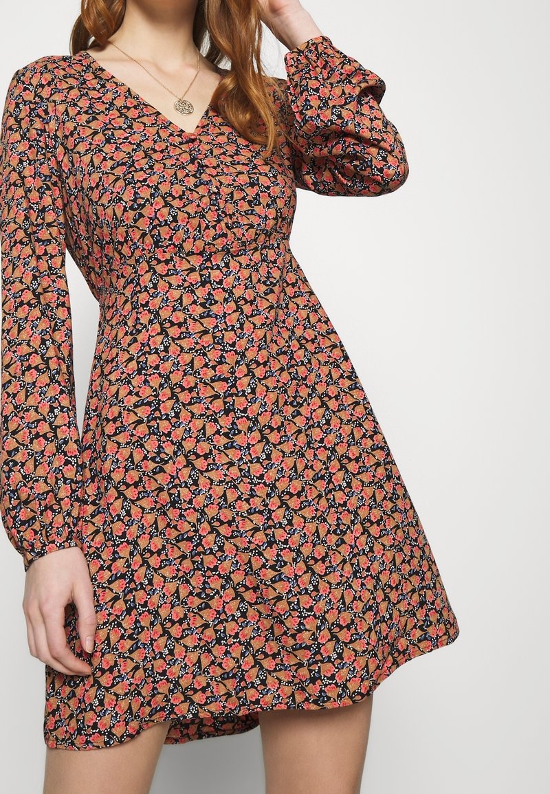 Femme portant une robe à manches longues à motifs floraux avec des fleurs roses et orange sur fond noir, debout devant un fond uni.