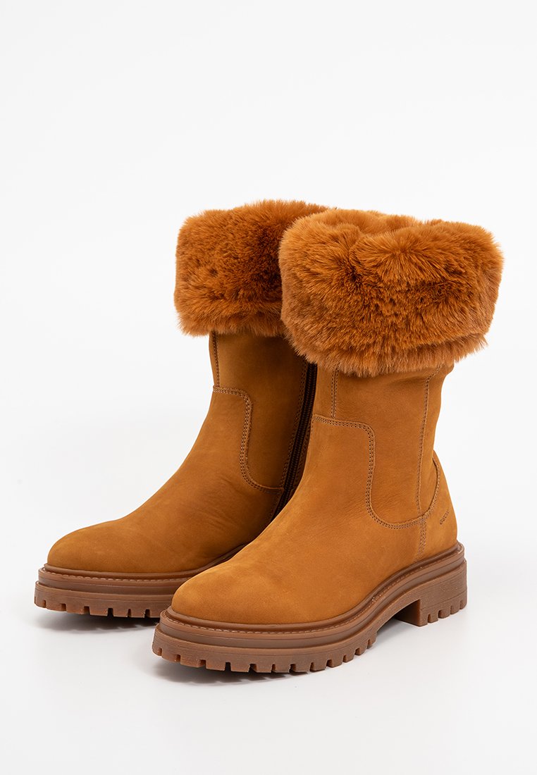 Botas de cuero color marrón claro hasta la mitad de la pantorrilla con suelas de goma marrón gruesas y puños de pelaje suave en color marrón anaranjado doblados, cremalleras laterales visibles.