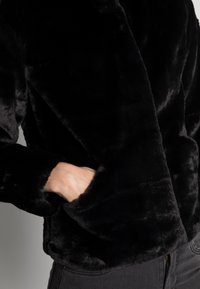 Veste en fausse fourrure noire avec une texture douce, dotée d'un large col et d'une poche sur le côté gauche. Le tissu a une finition lisse et brillante.