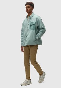Lichtblauwe canvas jas met een borstzak, gecombineerd met beige broek en grijze sneakers. Model staat met handen in de zakken tegen een witte achtergrond.