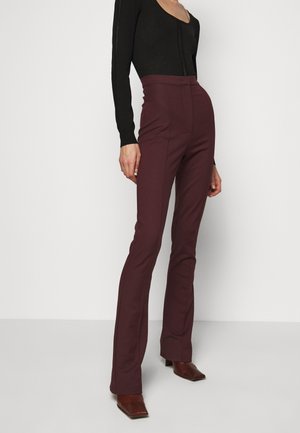 Henkilö, jolla on päällään tumman violetit korkea vyötäröiset suoralahkeiset housut, musta istuva pitkähihainen paita ja ruskeat teräväkärkiset korkokengät, seisoo valkoista taustaa vasten.