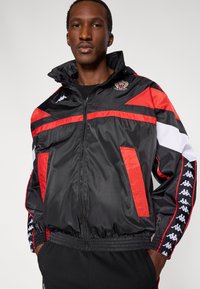 Czarna kurtka wiatrówka z czerwonymi i białymi akcentami, z przodu na zamek, z elastycznymi mankietami oraz naszywką z logo na piersi. Posiada boczne kieszenie.