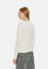Maglione di col beige chiaro con maniche lunghe, polsini e orlo a costine. Indossato con jeans grigi, che mettono in evidenza un design semplice e aderente.