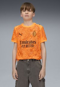 Maglia sportiva arancione con design marmorizzato, loghi e scritte nere. Maniche corte e scollo rotondo. Indossata con pantaloni cargo grigi.