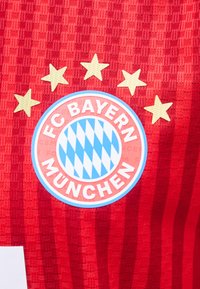 Λογότυπο του ποδοσφαιρικού συλλόγου FC Bayern München με πέντε χρυσά αστέρια πάνω από αυτό σε μια κόκκινη αθλητική φανέλα με υφή.