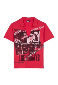 Polo shirt rouge à manches courtes avec un imprimé graphique comportant du texte et des images liées à une tournée, affichant une coupe décontractée et un col.
