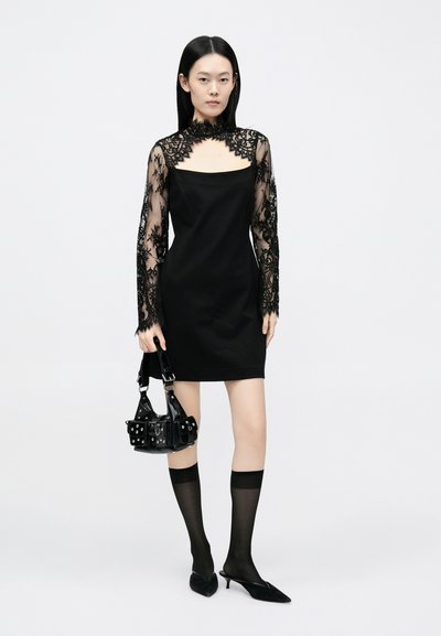 Svart kjole med lange, gjennomsiktige blondermer, firkantet utringning og en figursydd silhuett. Tilbehør inkluderer en liten svart veske og knelengde strømper.