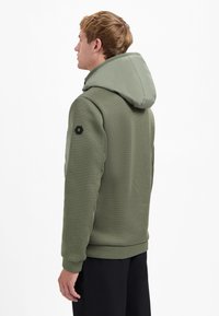 Groene hoodie met textuurstof, geribbelde manchetten en een logo patch op de linkerarm. Zichtbaar capuchondetail aan de achterkant.
