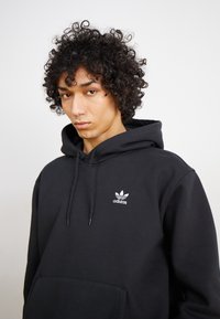 Μαύρος φούτερ με κουκούλα από βαμβάκι, με μπροστινή τσέπη και κορδόνι. Φέρει ένα λευκό λογότυπο της adidas κεντημένο στο στήθος. Στις πρώτες ειδήσεις υπάρχει κατσαρό μαλλί.