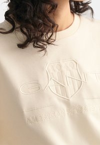 Beige sweatshirt med præget tekstlogo og skjolddesign. Blødt materiale med rund hals og ribstrikkede manchetter. Elegant, minimalistisk look.