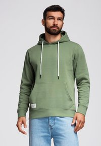 Sudadera de algodón verde con un bolsillo frontal, cordones ajustables y una etiqueta en el dobladillo. Combinada con jeans azul claro.