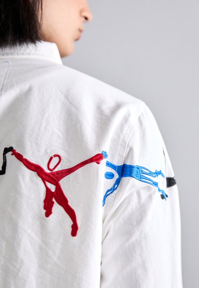 White shirt featuring colorful embroidery of figures in red, blue, and black. Soft texture with a classic collar and relaxed fit design.