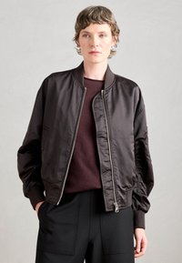 Black bomber jacket in satin material, featuring a zip front, ribbed cuffs, and side pockets. Worn over a burgundy sweater.