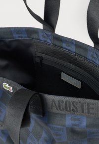 Torba na zakupy z czarno-niebieskim wzorem w kratę, z logo Lacoste, wewnętrzną siateczkową kieszenią na zamek oraz czarnymi materiałowymi uchwytami.