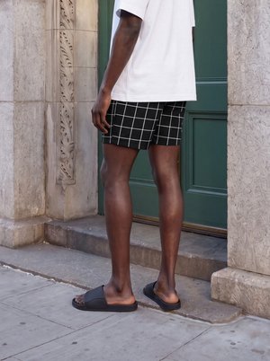Person trägt schwarze Slides und schwarze Shorts mit weißem Karomuster, steht auf Steinstufen vor einer grünen Tür.