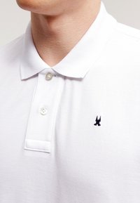 Närbild av en person som bär en vit polo-skjorta med två knappar knäppta och en liten svart broderad logotyp på bröstet.