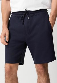Pantalones cortos deportivos color azul marino hechos de tela suave, con cintura elástica y cordón ajustable, y bolsillos laterales. Textura suave y longitud a la rodilla.