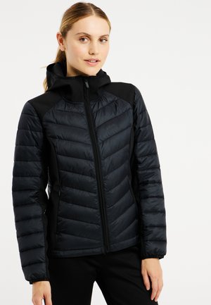 Jonge vrouw draagt een zwarte gewatteerde jas met ritssluiting en capuchon, staande tegen een effen lichte achtergrond met een lichte glimlach.