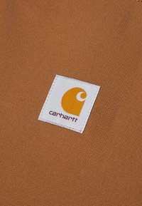 Smeđi platneni materijal s bijelom etiketa sa šavom koja sadrži okrugli narančasti logo i tekst "carhartt" u tamnocrvenoj boji.